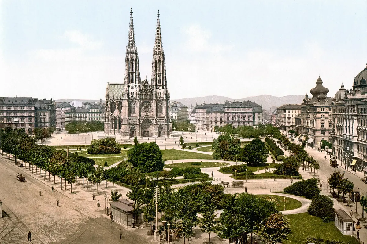 Una stampa a colori del 1900 circa che mostra la Votivkirche dalle due torri attraverso una piazza aperta con alberi, sentieri e alti edifici cittadini.