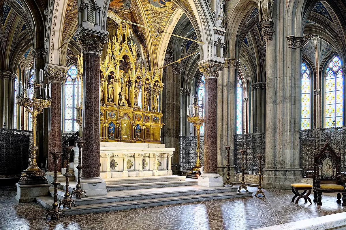 L'altare maggiore dorato della Votivkirche su gradini di marmo, incorniciato da colonne in granito rosso, con un baldacchino in oro intagliato e vetrate istoriate sullo sfondo.
