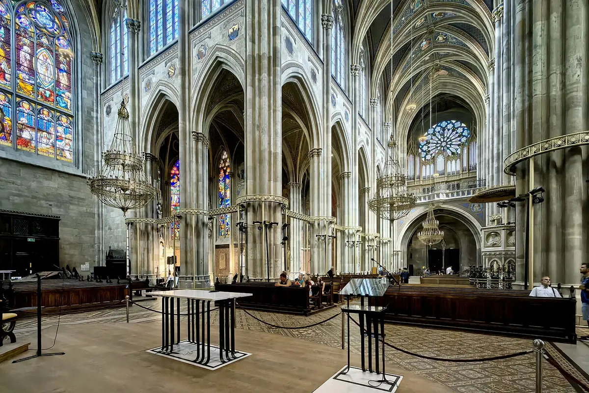 La navata della Votivkirche in luce ordinaria, con alti archi a sesto acuto, pilastri in pietra grigia, una vetrata blu e rossa a sinistra e il presbiterio in lontananza.