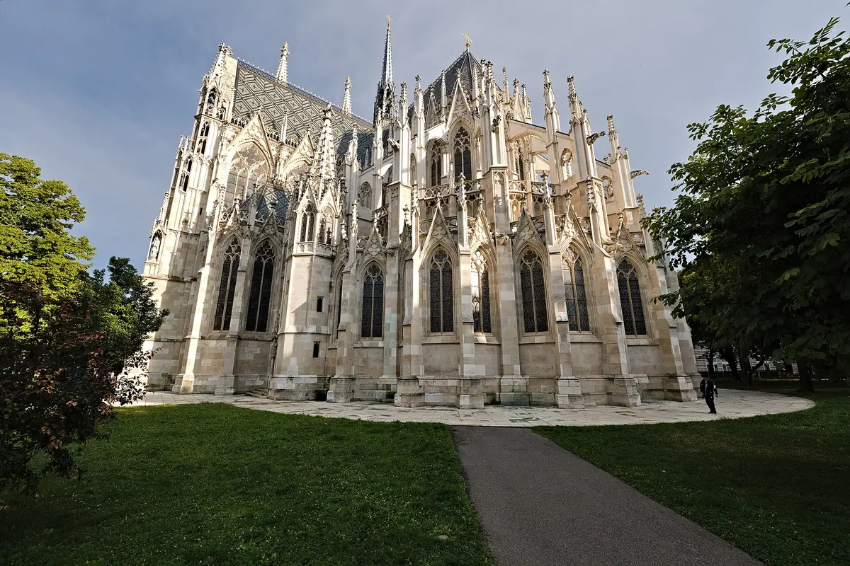 L'estremità orientale della Votivkirche vista dal parco circostante, con alte finestre, contrafforti e pinnacoli in pietra chiara sopra un prato verde.
