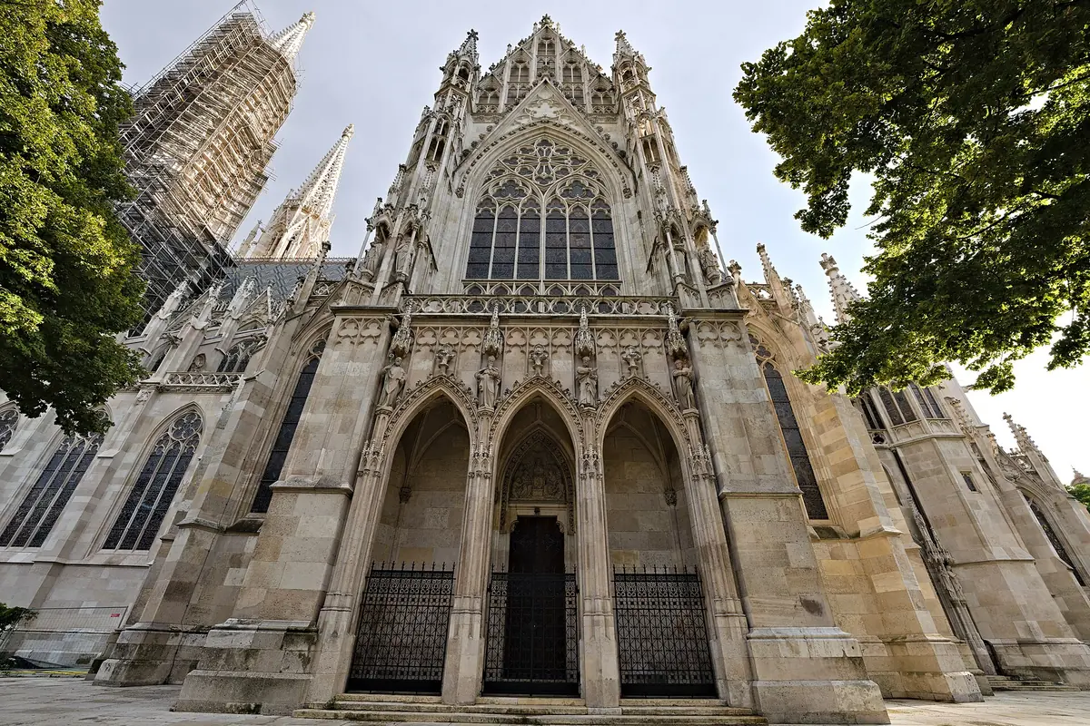 Il portale principale della Votivkirche, tre ingressi ad arco a sesto acuto sotto un grande finestrone traforato, con impalcature su una torre a sinistra.