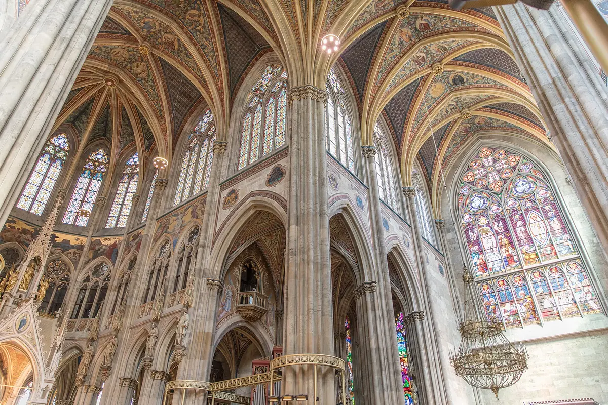 Sguardo verso l'alto all'interno della Votivkirche: volte a costoloni color crema, alte finestre a sesto acuto e un grande rosone istoriato sulla destra.
