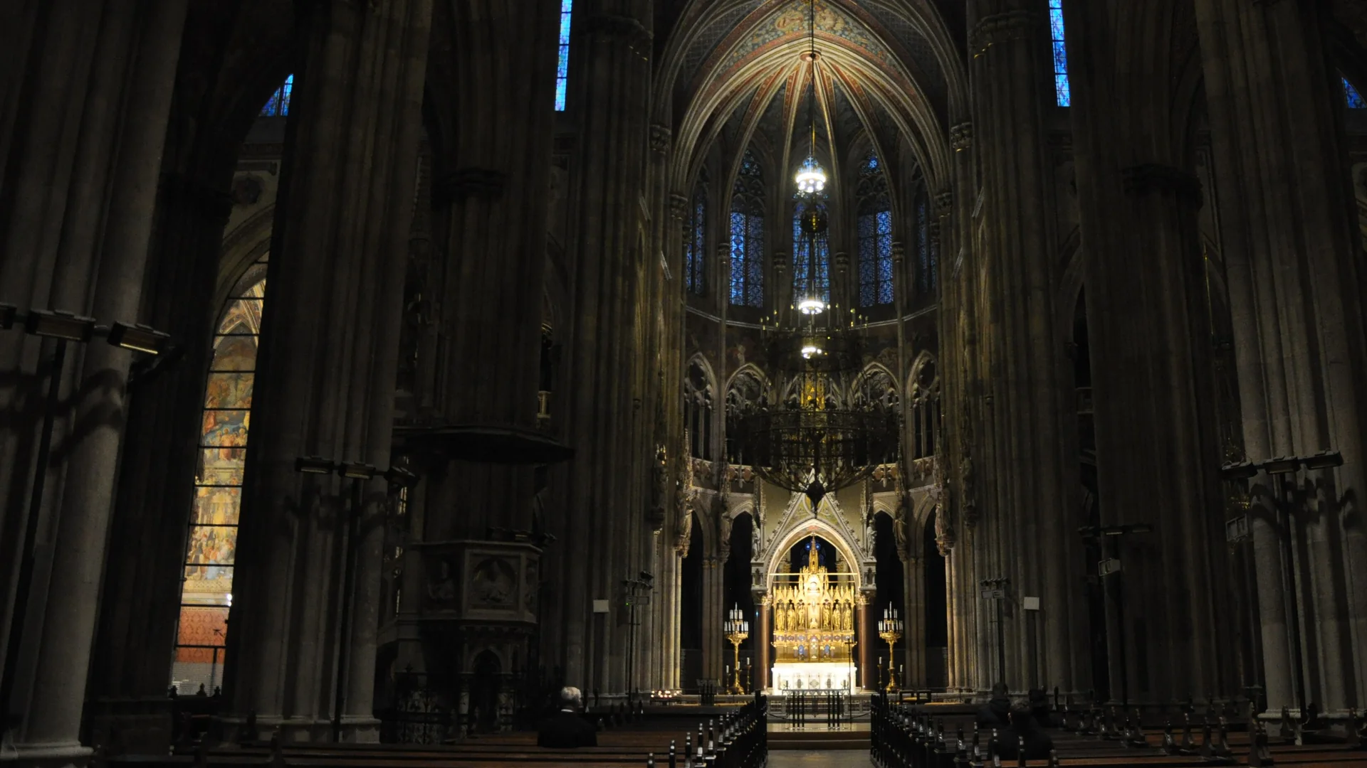 La navata della Votivkirche in luce ordinaria: un corridoio buio tra alte pilastri in pietra grigia conduce a un altare dorato, con vetrate blu e una volta dipinta in alto.
